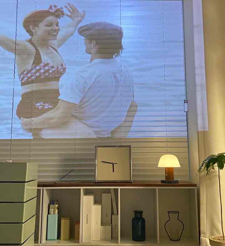 白と木目調が調和した本棚とテーブルランプのある雰囲気の良いリビング、ブラインドに大きなウォールデコが投影されている