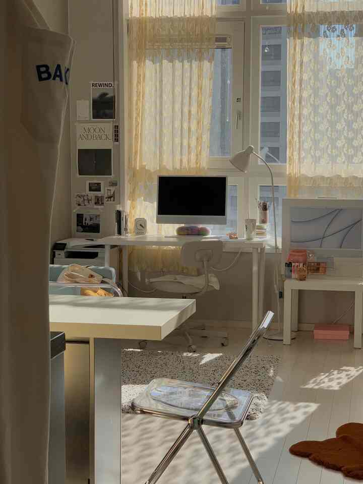 暖かいベージュのカーテンと白い壁、床、家具が調和するホームオフィス空間、中央に広いデスクと透明な椅子が配置されている