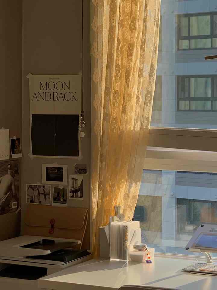 黄色のレースカーテンとモノトーンの壁、ポスターが配置された窓際の居心地の良いホームオフィス空間