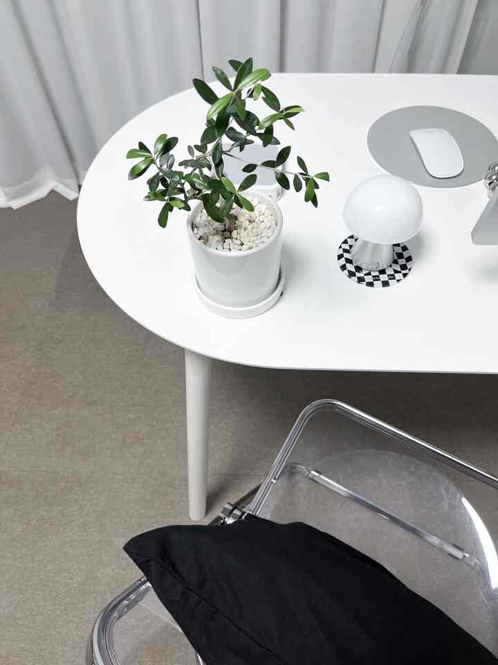 ホワイトトーンのデスクと透明チェアがある空間に、中央に緑のオリーブの木の鉢植えとワイヤレスLEDマッシュルームランプが配置されたすっきりとしたミニマルインテリア