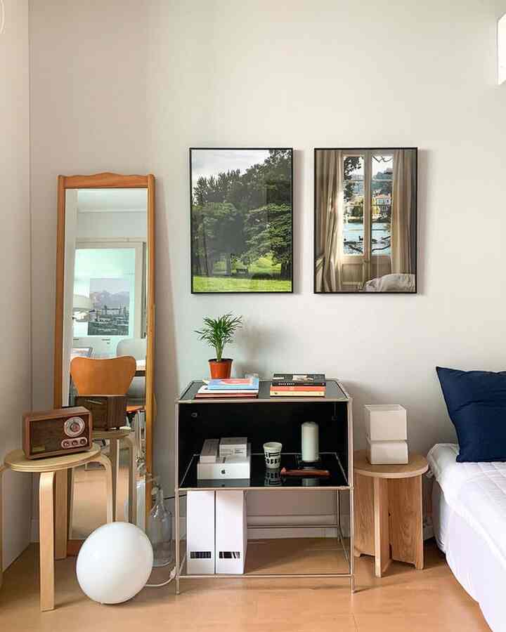 白い壁と木目調の床や家具がある寝室、スツールの上のヴィンテージラジオと大型の鏡、壁に掛けられた風景のポスターが調和したシンプルなインテリア
