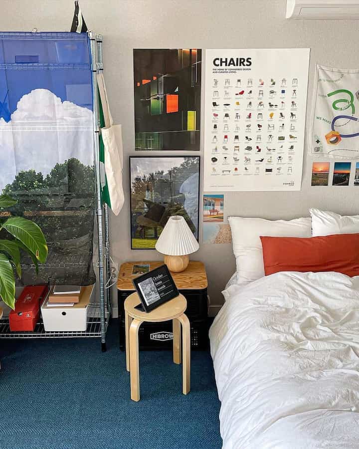 ネイビートーンのラグが敷かれた寝室空間、木製スツールと多様なポスター、白い寝具が調和するナチュラルモダンなインテリア