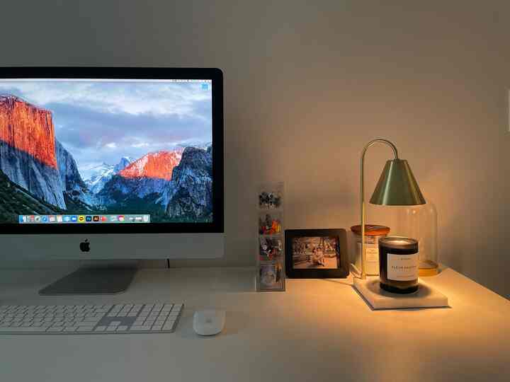 白とシルバーのホームオフィスデスク、灯されたキャンドルとモダンなコンピューターが配置された温かみのある作業空間