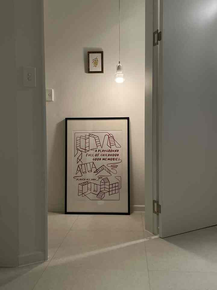 ホワイトトーンの玄関空間、床に置かれた額縁と上部のポイント照明が配置されたシンプルで清潔感のあるインテリア