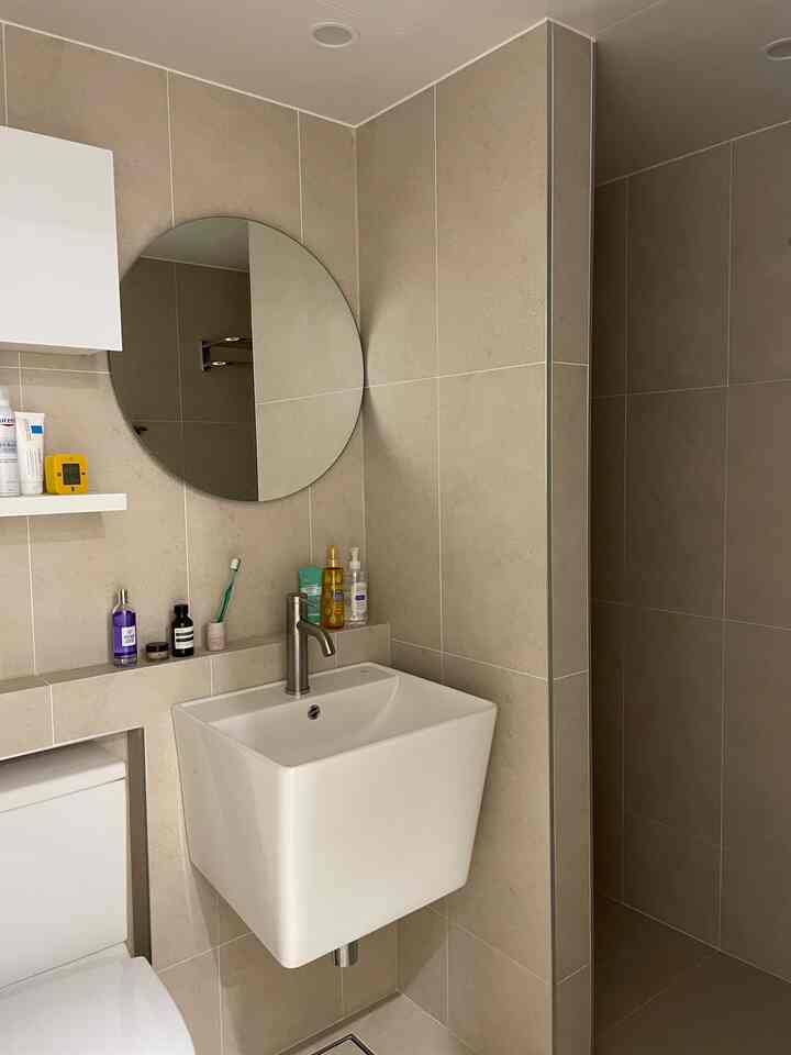 ベージュ色のポーセリンタイル壁と塀壁があるシンプルなバスルーム、丸い鏡と洗面台が配置された落ち着いた空間