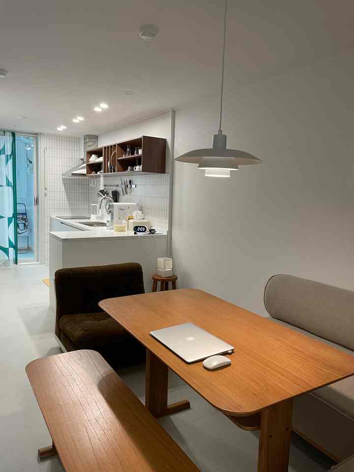 白い壁と木目調の家具が調和するキッチンとリビング、ペンダントライトの下に大きなテーブルとアームチェアが配置された温かみのある空間