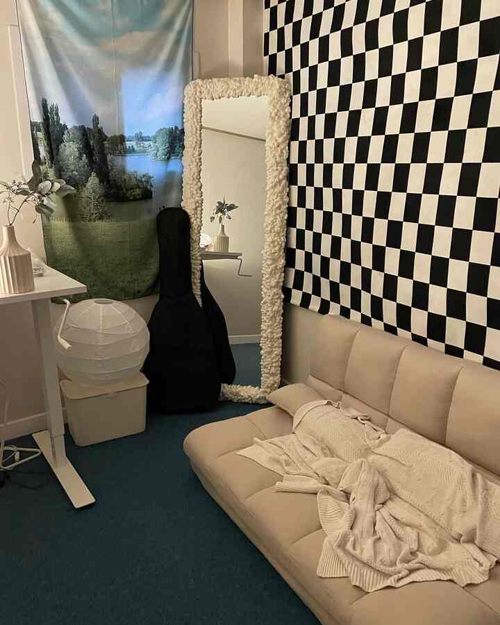 暗いブルーのラグが敷かれた狭いリビング、ベージュのソファベッドとチェッカーボード柄の壁カバー、全身鏡が配置された雰囲気のある空間