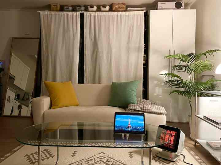 明るいホワイトトーンのリビング、ベージュの2人掛けソファに黄色とミント色のクッション、ガラスのコーヒーテーブルと室内植物が配置された雰囲気のある空間