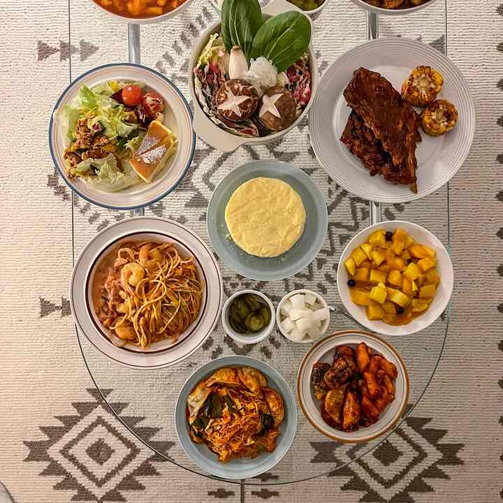 ベージュ調のラグの上に丸いガラスのダイニングテーブルが置かれ、多様な皿やボウルに料理が盛り付けられたダイニングスペース