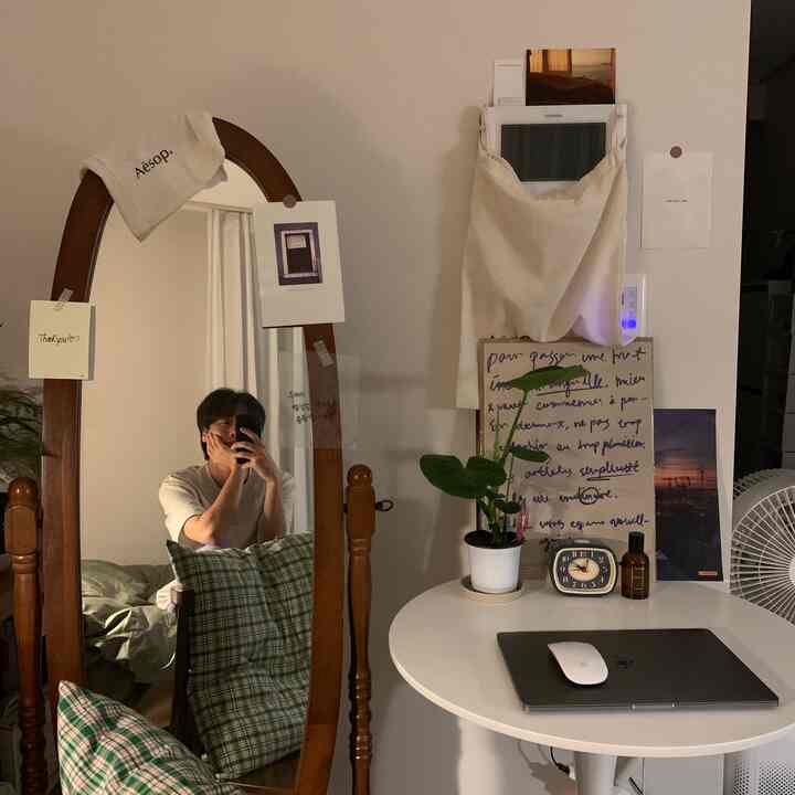 白い壁と木製スタンドミラーがあるワンルーム寝室、丸い白いテーブルにノートパソコン、目覚まし時計、植物、イソップのディフューザーが置かれた落ち着いたホームオフィスのインテリア