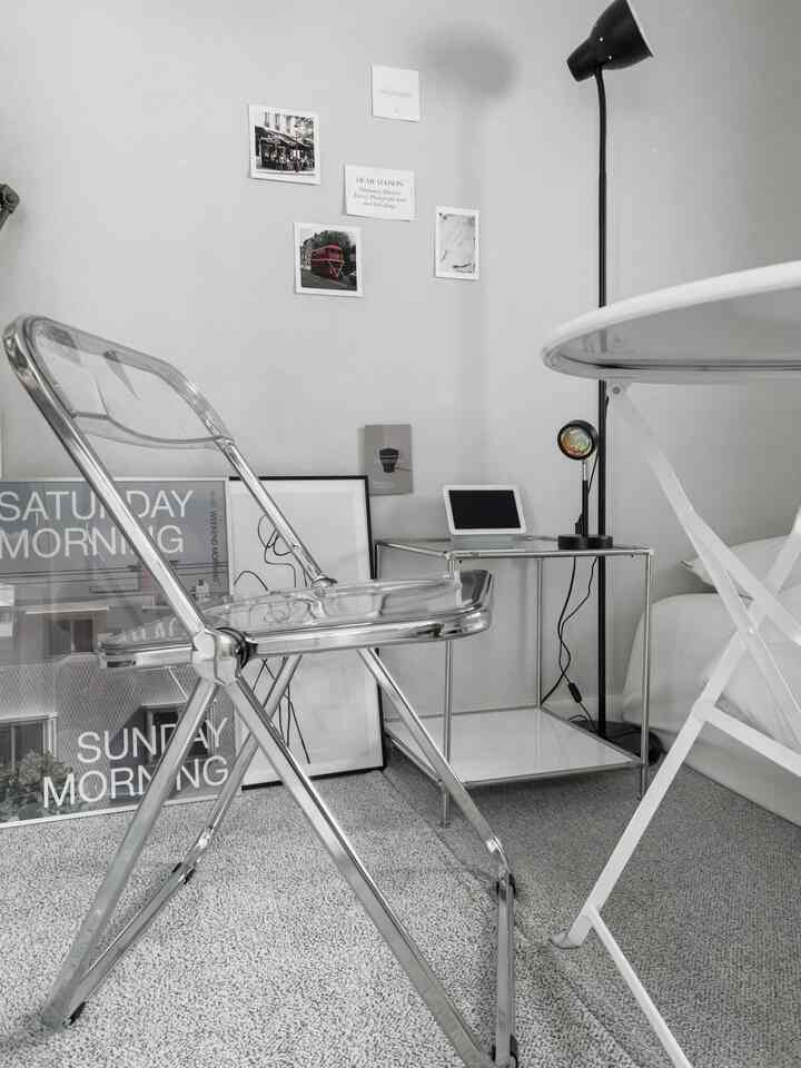 モノトーンとホワイトのワンルーム、透明なモジュール椅子と鉄製ナイトテーブル、ポスターが配置されたモダンでミニマルなインテリア