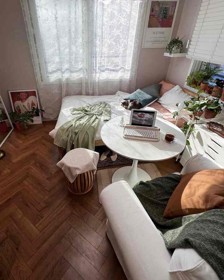 ピンクの壁紙とホワイト家具が調和した寝室兼リビング、丸テーブルとソファ、植物が配置された明るい空間