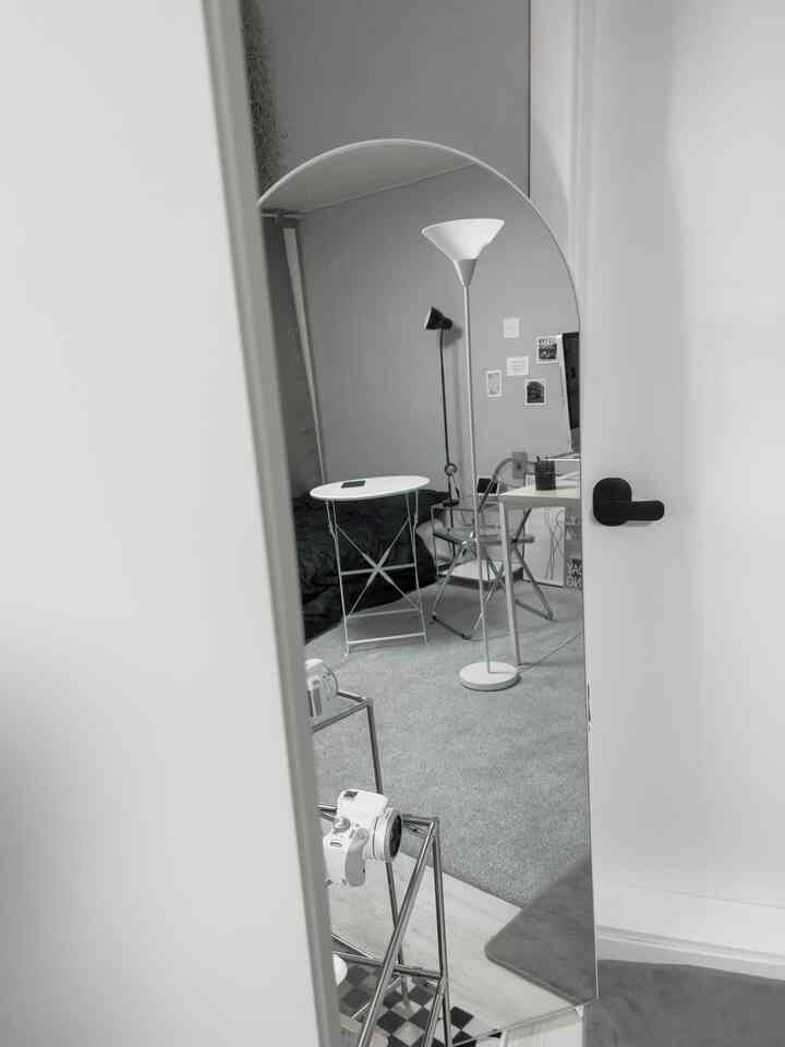 モノトーンでモダンなワンルーム、アーチ型の大きな鏡とフロアランプ、白いデスクと黒いベッドが配置された清潔感のあるインテリア
