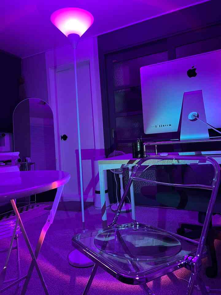 紫色の照明が照らすワンルーム空間、透明な椅子と白いデスクが配置されたモダンなホームオフィスのインテリア