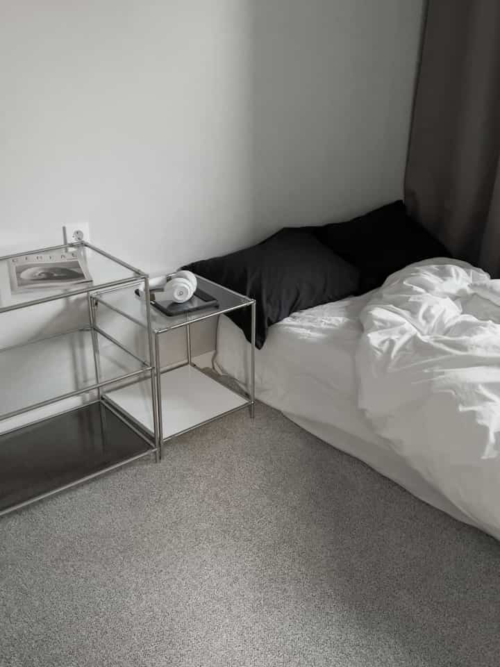 モノトーンの小さなワンルーム寝室、白い寝具と黒い枕のベッドと金属製ナイトテーブルが配置されたシンプルでモダンなインテリア