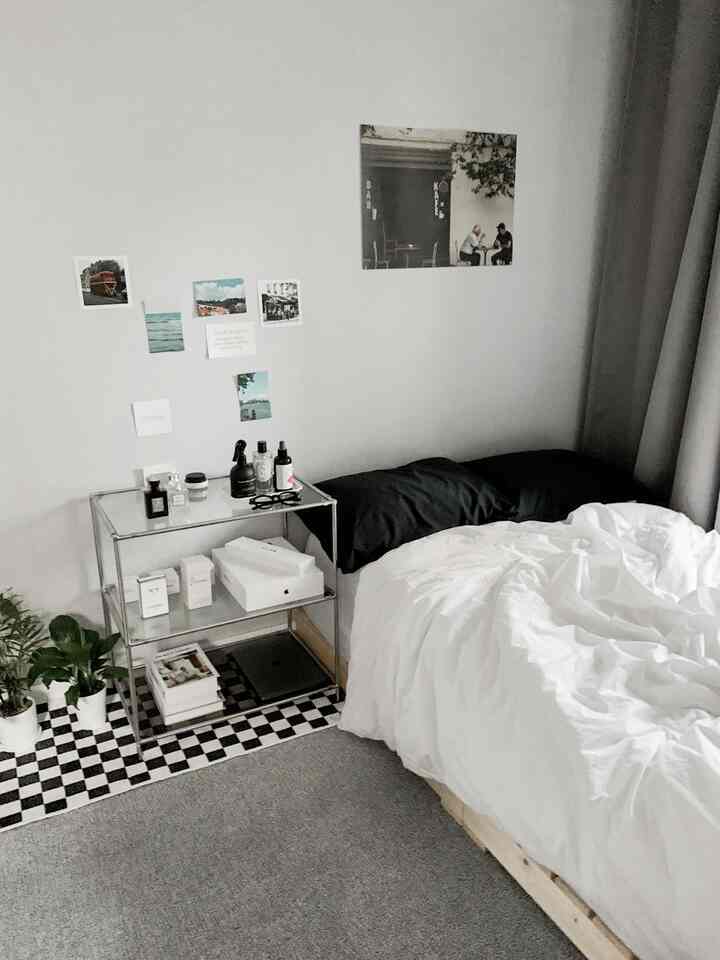 モノトーンのモダンスタイルワンルームの寝室、黒い枕と白い掛け布団カバーのベッド、チェッカーボードのラグ、金属製の収納棚、壁にポスターが配置されたすっきりした空間