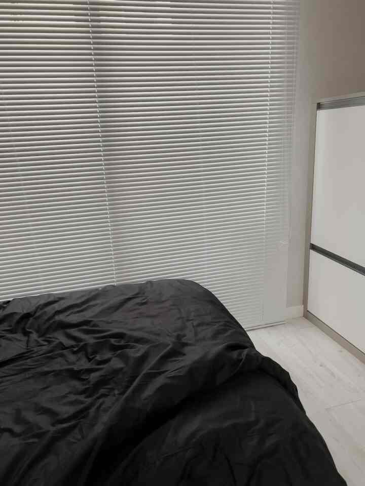 白を基調としたワンルームの寝室、ブラインドと黒い寝具が調和したモダンで清潔感のある空間