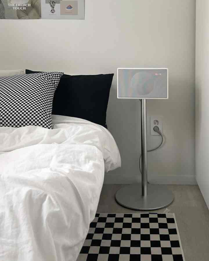 白い寝具とモノトーンのチェッカーパターンの枕がある寝室、ジェネバのスピーカーがスタンドに置かれたすっきりとした空間