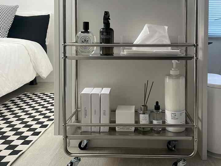 モノトーンと白、ウッドトーンが調和した寝室、チェッカーボードの足マットとミニマルな金属収納カートが配置された雰囲気のあるインテリア。