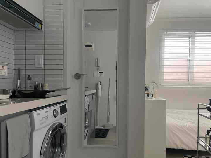 白を基調とした細長いキッチンとトイレの空間、壁掛け鏡と木製ブラインドが配置された清潔感のあるインテリア