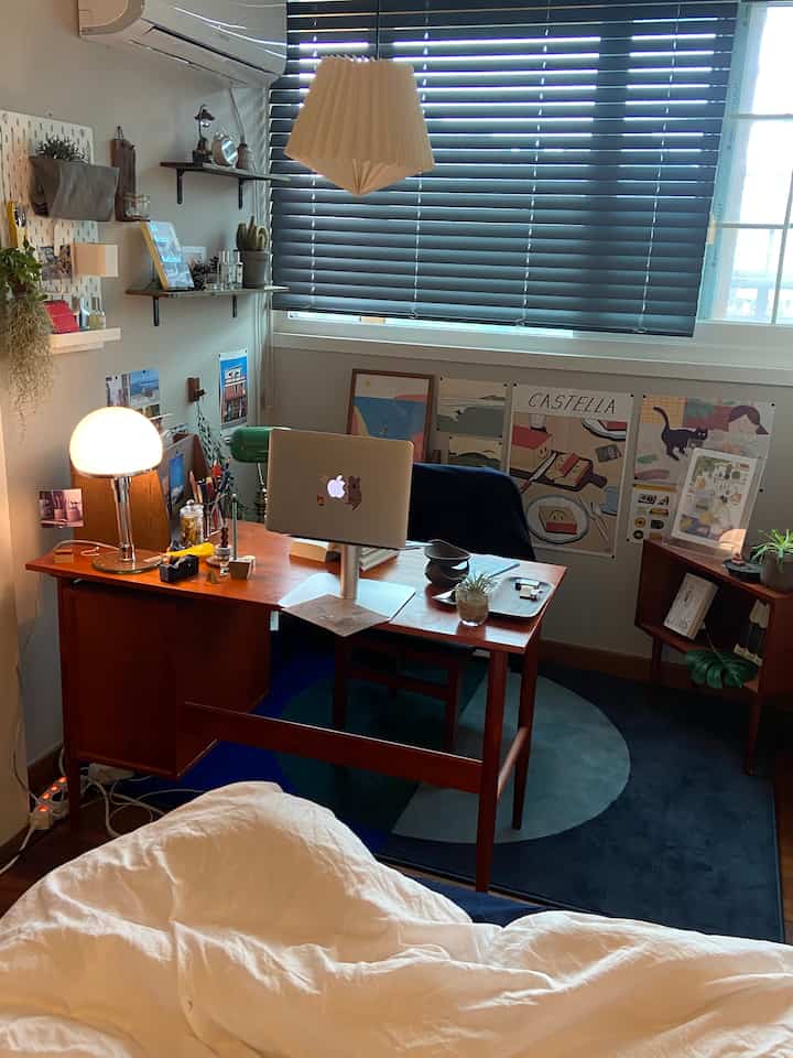 ネイビーブラインドと木製デスクが配置されたコンパクトなホームオフィス、ホワイトの寝具が調和する雰囲気のある空間