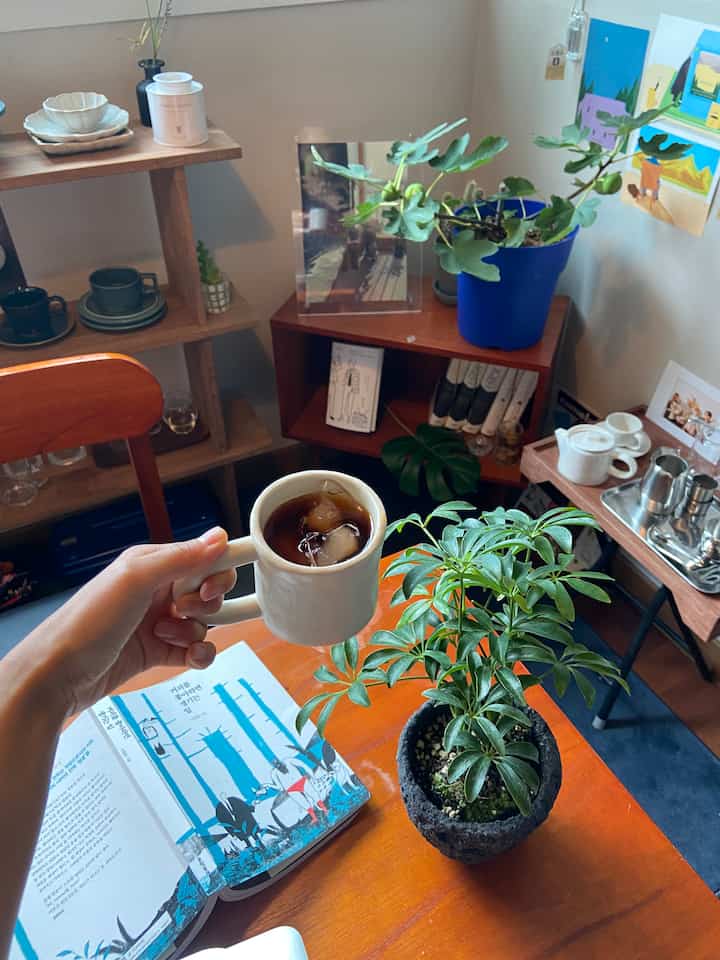 温かみのある木目調のコンパクトな6畳の部屋にあるおうちカフェ、植物と本が置かれ、シンプルなテーブルにコーヒーマグが配置された落ち着いた雰囲気のインテリア