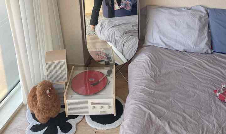 薄紫色の寝具が敷かれた寝室、ベッド横にベージュのナイトテーブルと赤いLPターンテーブル、全身鏡が配置された落ち着いた空間