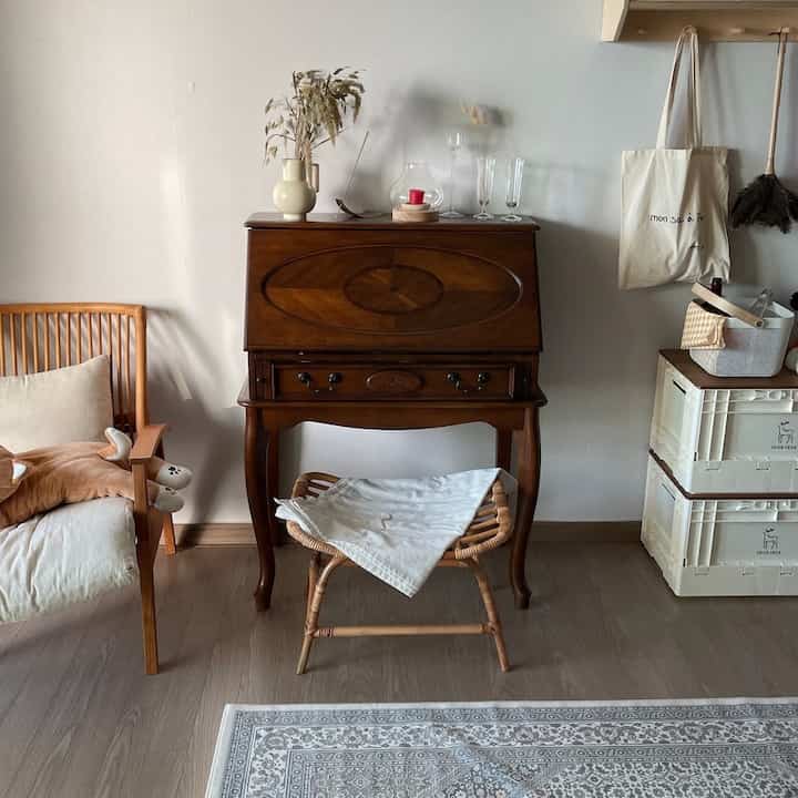 ナチュラルトーンのリビング、木製のセクレタリーデスクとラタンのスツール、クッション付きのアームチェアが配置された落ち着いた雰囲気のインテリア