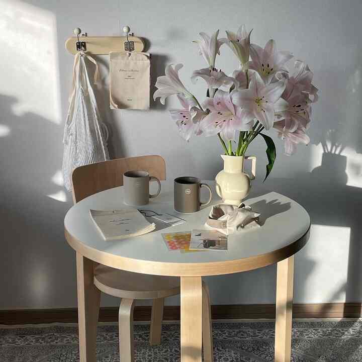 白とナチュラルトーンのワンルーム空間、丸い木製テーブルと椅子が配置され、花瓶とマグカップが置かれた雰囲気のあるホームカフェインテリア