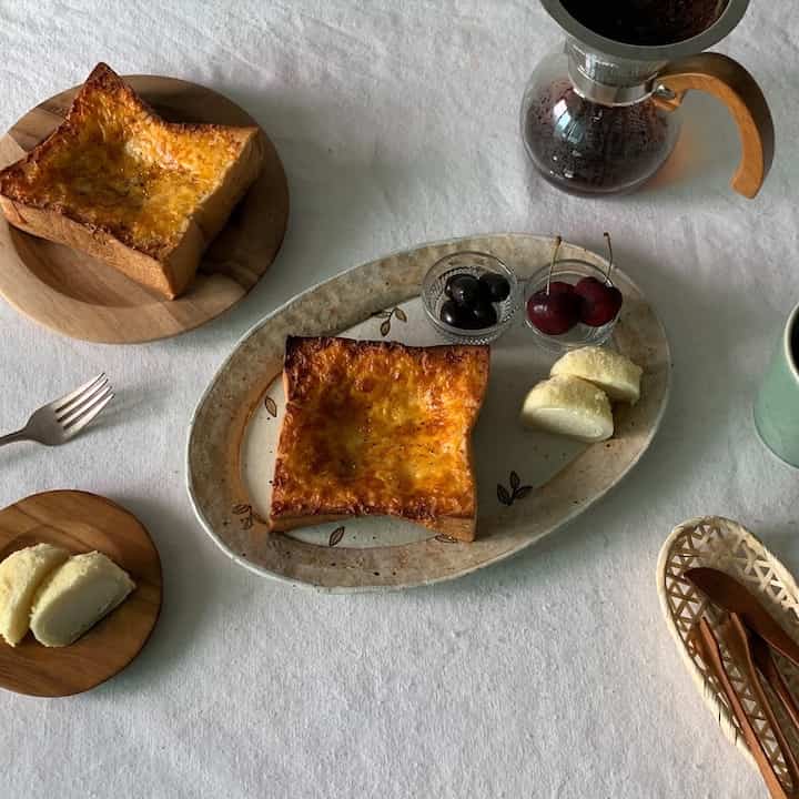ホワイトのテーブルクロスに木のトーンの皿とクラシックな陶器トレイが配置され、おうちカフェスタイルのコーヒーとトーストがあるミニマルで落ち着いた空間
