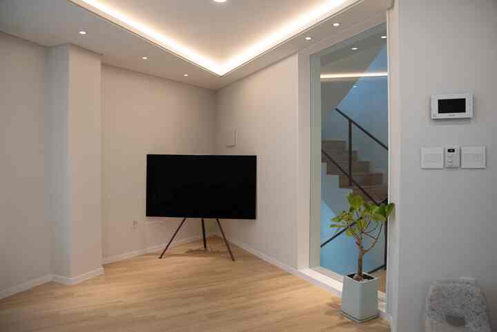 白と木目調が調和したミニマルなリビング、三脚型のSamsungテレビと階段近くの小さな植物が配置された清潔感のある空間
