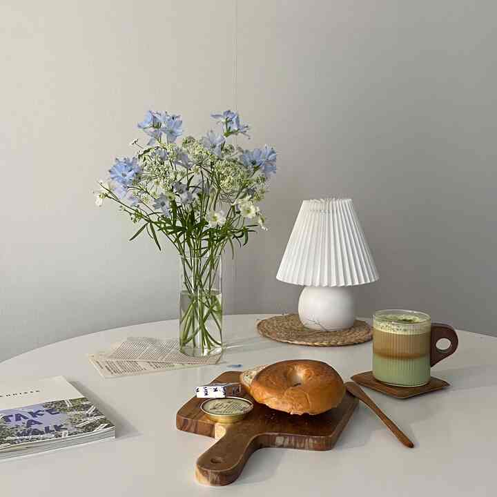 白と木目調のテーブルに花瓶、テーブルランプ、ホームカフェの飲み物とパンが配置されたシンプルでナチュラルな雰囲気のインテリア