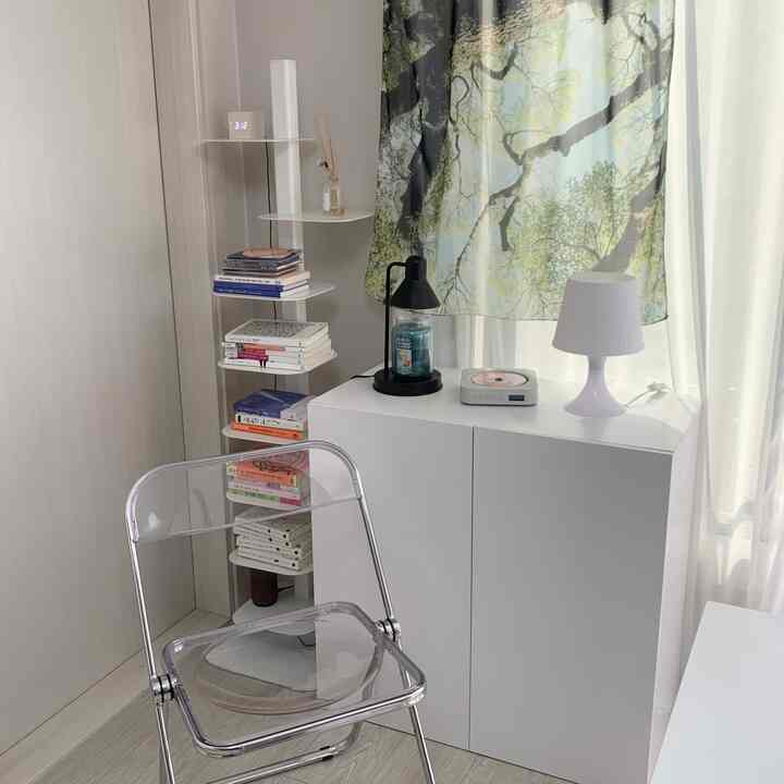 ホワイトとナチュラルカラーの寝室コーナー、透明な折りたたみ椅子と本棚、テーブルランプが配置された清潔感のあるインテリア