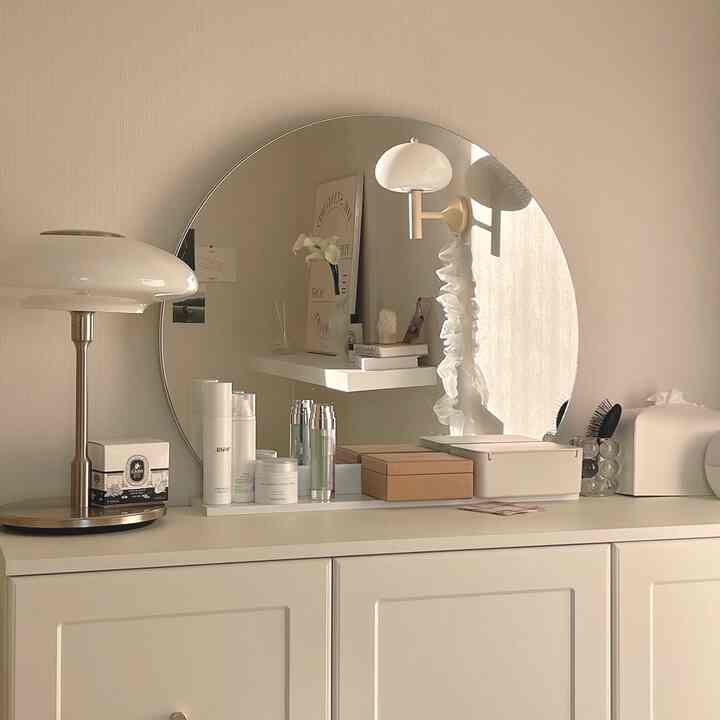 ホワイトとベージュのトーンの化粧台空間、大きな丸い鏡とテーブルランプが配置されたシンプルで清潔感のあるインテリア