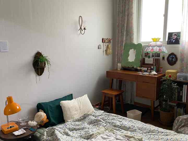 ナチュラルな木調家具とヴィンテージの花柄カーテンが配された、化粧台と壁付け照明のある居心地の良い一人暮らしの寝室