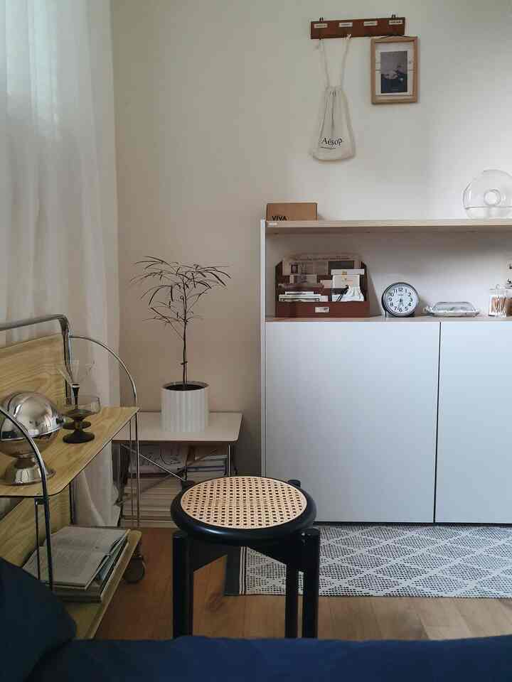 ナチュラルカラーとホワイトのリビング、シンプルな収納キャビネットと木目調の家具、小さな植物とスツールが配置された落ち着いた空間