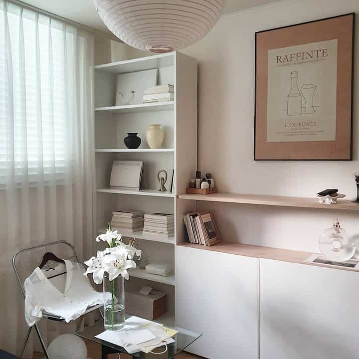 白とナチュラルな色調のリビングルーム、書棚、額縁、カーテン、ガラステーブルが配置された落ち着いた印象の空間