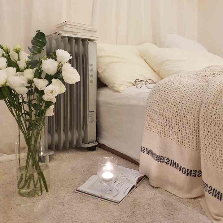 明るいホワイトとベージュのトーンの小さな寝室、ベッドと花瓶、オイルヒーターが温かみのある雰囲気を作り出すインテリア