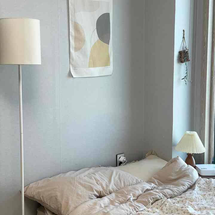 柔らかいベージュ色の寝室、左にフロアランプ、右にテーブルランプ、中央の壁に抽象的なポスターが配置された雰囲気のある空間