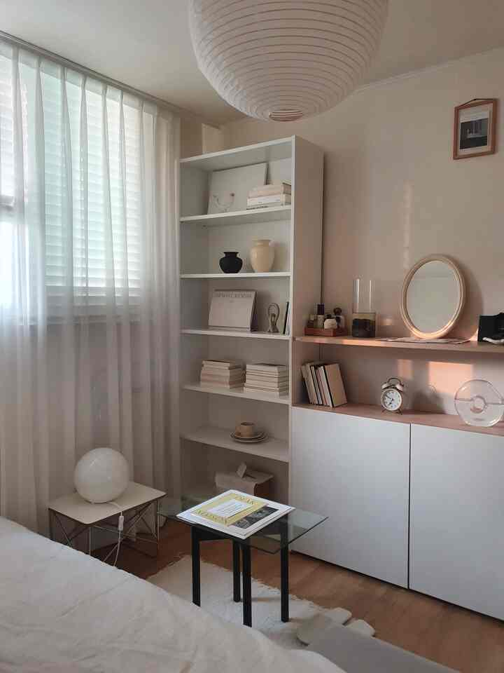ホワイトとベージュの色調の寝室、ミニマルな雰囲気のある本棚とキャビネットが配置された空間