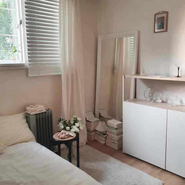 白とベージュのトーンの寝室、窓辺にカーテンと鏡、積まれた本とスツールが配置された落ち着いた雰囲気の空間