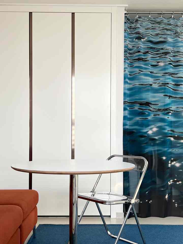 白と青のトーンのワンルーム、透明な椅子と円形テーブル、オレンジ色のソファが配置された印象的なインテリア