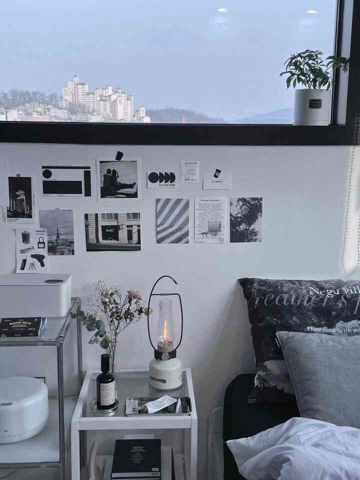 白と黒のトーンの寝室空間、ナイトテーブルにはムードランプとディフューザーが置かれ、窓際には小さな植物が配された清潔感のあるインテリア