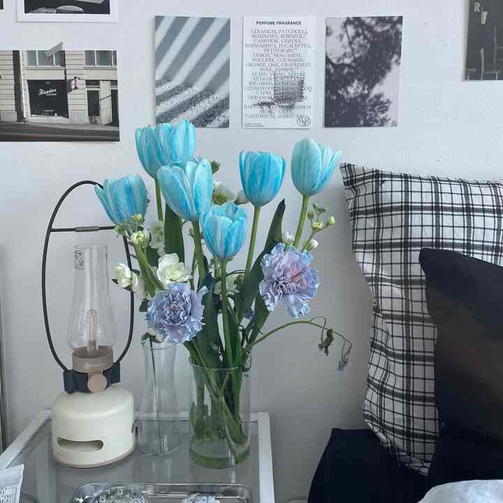 モノトーンのベッドルームの隅に、青いチューリップと紫のカーネーションが入った透明なガラスの花瓶が置かれた小さなサイドテーブルが配され、落ち着いた雰囲気のあるインテリア