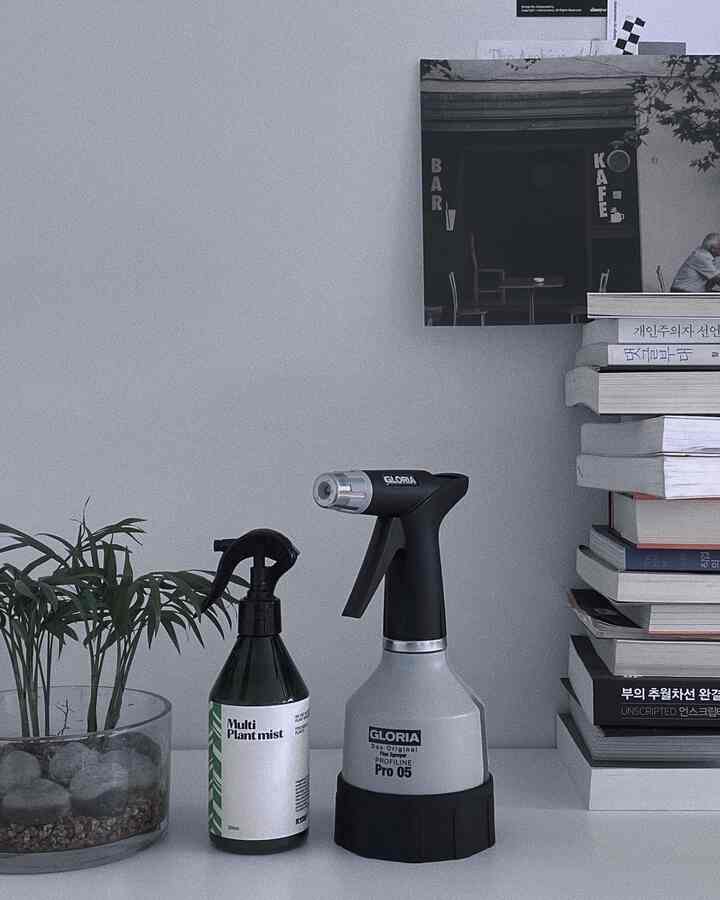 グレーとホワイトの色調の寝室のテーブル上に、スプレーボトルと透明な植物の鉢、整頓された本が置かれたモダンなインテリア