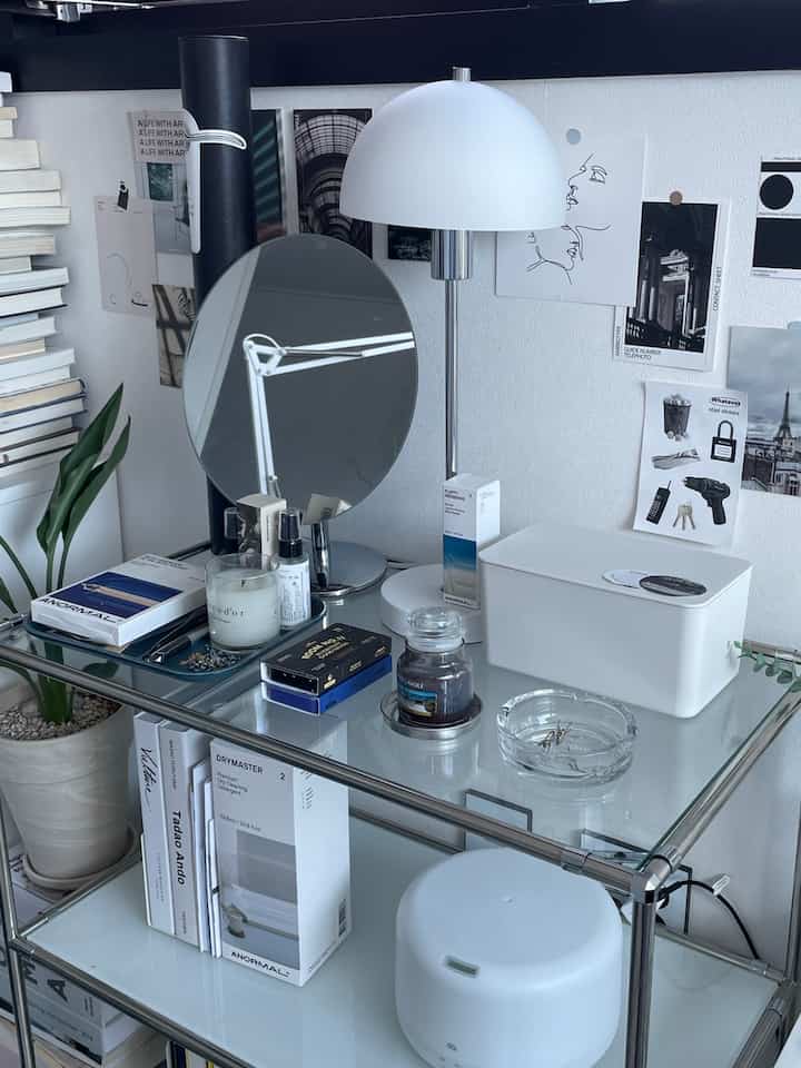 白と透明なトーンの本棚とガラステーブルが配置されたミニマルな作業空間、テーブルランプと丸い鏡が印象的なインテリア