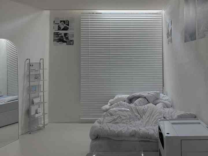 白を基調とした狭くて変形間取りの寝室、白いブラインドと布団、鏡が配置されたシンプルなインテリア