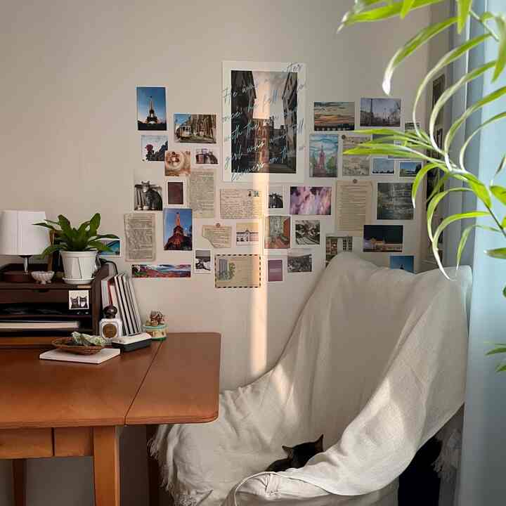 暖かい木目調と白のトーンの小さな部屋、ポストカードの壁飾りと木製の机や植物が配された雰囲気のある書斎