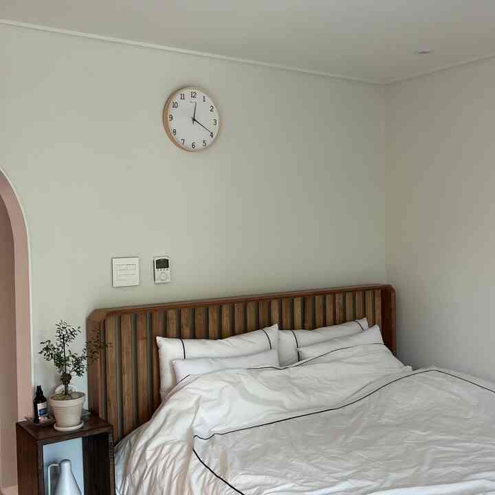 白と茶色のトーンの寝室、木製ベッドフレームと壁掛け時計、ナイトテーブルが配置された清潔で自然な雰囲気のあるインテリア