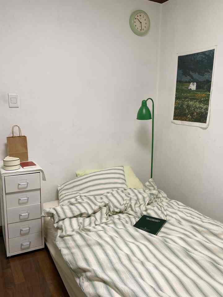 白い壁とナチュラルな木目調の床、緑のフロアランプと時計がある6畳の寝室、ストライプの掛け布団が配置されたシンプルで温かみのあるインテリア